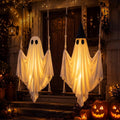 Two glowing white ghosts in witch hats hanging on rope swings in front of a porch at night