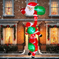 Santa inflatable hanging from a window holding a Merry Christmas sign with ornaments below on a brick house